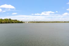 A serene lake view featuring green trees and distant sailboats under a blue sky.
