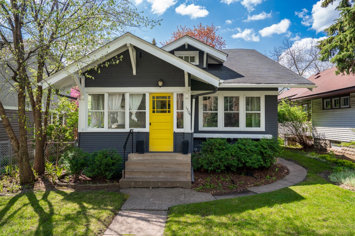 Charming bungalow with vibrant yellow door welcomes guests in a peaceful residential neighborhood surrounded by mature trees and spring blooms.
