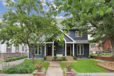 A two-story dark blue house with white trim, surrounded by greenery and pink flowers in pots.