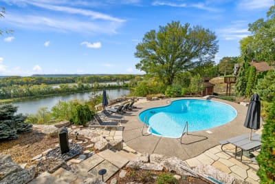 A swimming pool surrounded by lounge chairs, with a river and trees in the background under a blue sky.