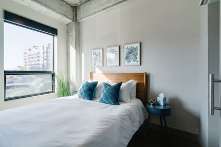 A stylish bedroom with white bedding, turquoise pillows, abstract art, and a window overlooking an urban landscape.