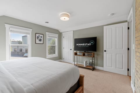 Serene bedroom with a white bed, flat-screen TV, and natural light from windows.