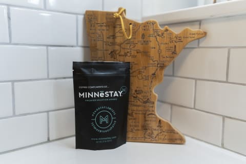 Black coffee bag next to a wooden cutting board shaped like Minnesota on a white countertop.