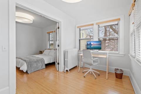 A well-lit bedroom featuring a bed, a workspace with a monitor, and large windows.