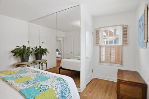 A cozy bedroom featuring a patchwork quilt, mirrored wardrobes, and warm wooden floors with a window and plants.