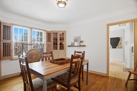 A cozy dining area with a wooden table and chairs near large windows, featuring a decorative shelf and glass cabinet.