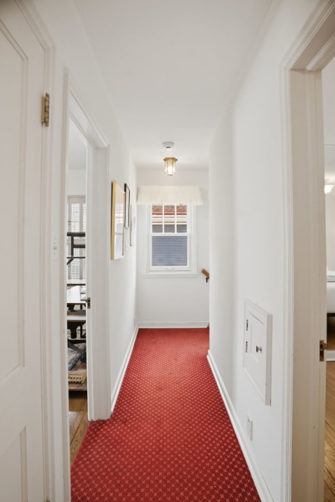 A bright hallway with white walls and a red carpet, featuring a window and light fixture.