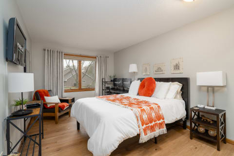 A modern bedroom featuring a large bed with white linens, an orange throw blanket, and a cozy reading nook.
