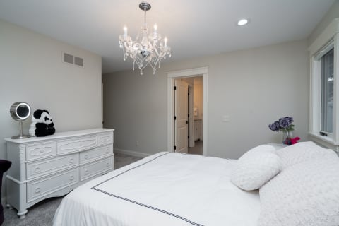 A cozy bedroom with a white bed, chandelier, and panda plushie on a dresser.
