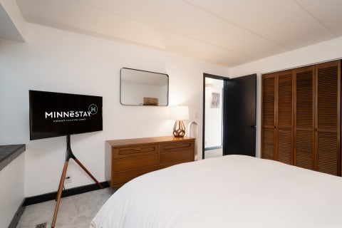 Modern bedroom interior with a television on a tripod stand, wooden dresser, and unique lamp.