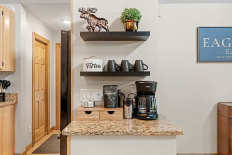 A kitchen corner with a coffee maker, mugs on a shelf, and decorative elements.