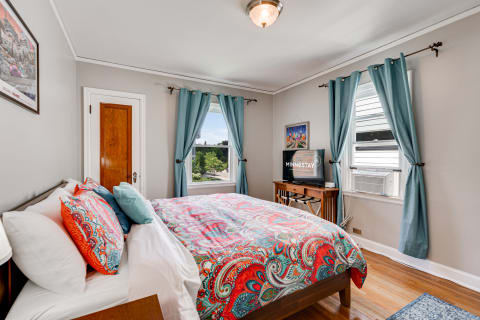 A cozy bedroom with a bed covered in colorful bedding, windows with aqua curtains, and a TV on a wooden desk.