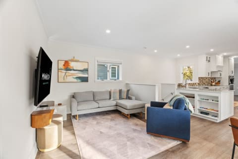 A bright open-plan living area with a gray sectional sofa, vibrant blue accent chair, abstract wall art, and modern kitchen in the background.
