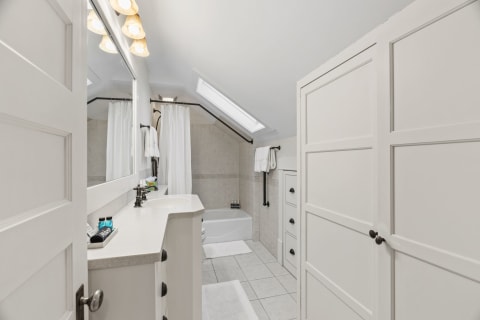 A bright and modern bathroom featuring a white vanity, a bathtub, and towels, illuminated by a skylight and pendant lights.