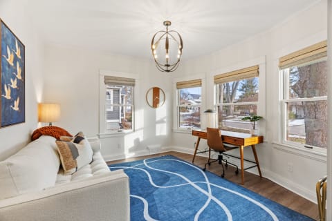 Bright living area featuring a white sofa, large windows, and a wooden desk with a modern chandelier.