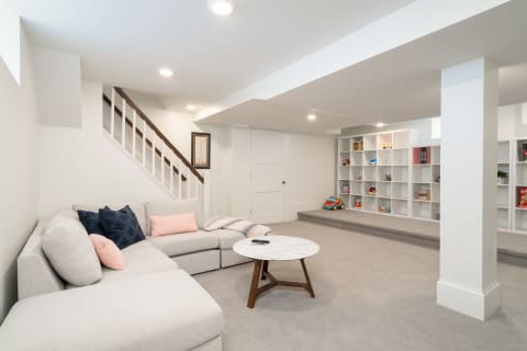 A bright basement with a sectional sofa, coffee table, and white shelving filled with toys and books.