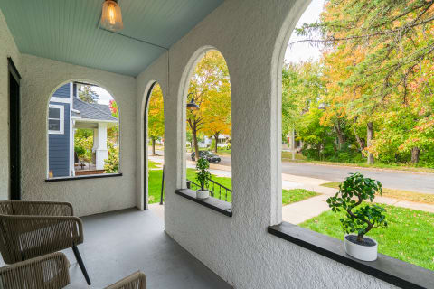 A porch with archways, rattan chairs, and a potted plant, revealing a view of autumn trees and a street.