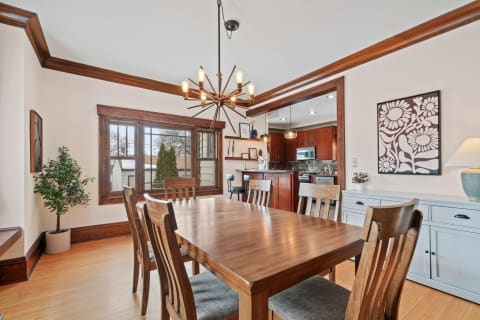 A dining room featuring a wooden table, chairs, and a modern chandelier with an adjoining kitchen visible.