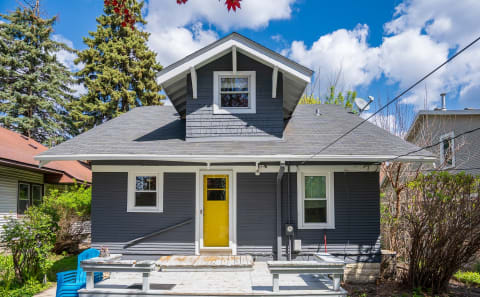 A gray house with a yellow door and blue chairs on the porch against a blue sky.