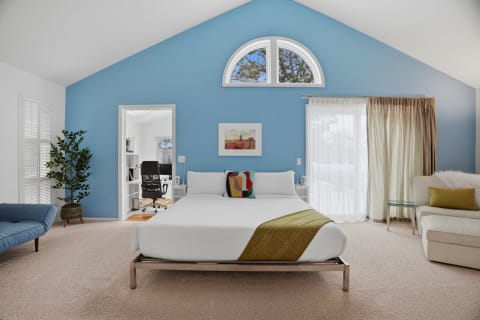 A bright bedroom featuring a vaulted ceiling, a white bed with a green throw, and a blue lounge chair, complemented by natural light and decor.