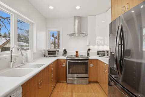 Modern kitchen with stainless steel appliances, wooden cabinetry, and large windows overlooking a snowy neighborhood.