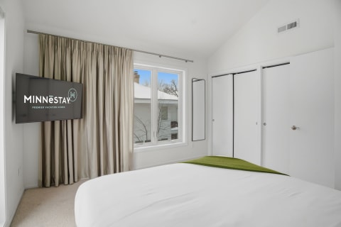 Modern bedroom featuring a queen bed, wall-mounted TV, and natural light from a large window.
