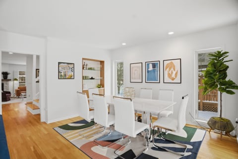 A bright dining room featuring a sleek white table, white chairs, colorful artworks, and a built-in shelf.