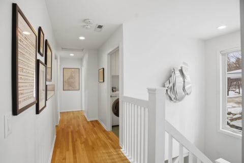 A bright hallway with wooden floors, framed artworks on the walls, and a window letting in natural light.