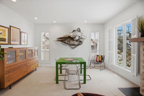 A bright room featuring a green table, modern chairs, and a metallic wall sculpture, illuminated by sunlight from large windows.