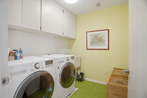 Modern laundry room featuring white washing machines, yellow walls, and wooden baskets on green carpet.