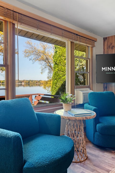 A living room with blue chairs, a round woven table, and a scenic view of a lake through large windows.