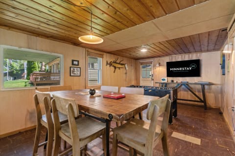 A rustic dining space with a large table, wooden chairs, and a foosball table, illuminated by pendant lighting and a view of the outdoors.