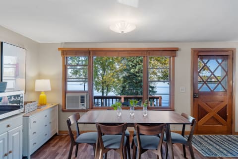 Cozy dining area with large windows overlooking a lake and trees, featuring a wooden table and stylish chairs.