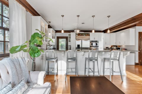 A modern kitchen with a white couch, bar stools, and stainless steel appliances illuminated by natural light.