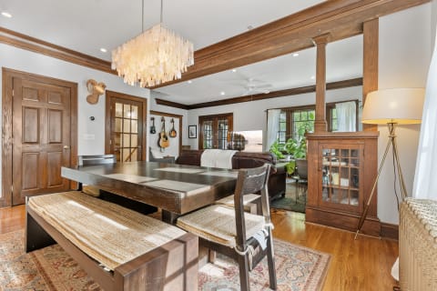 Modern dining room with a dark wood table and bench, layered chandelier, and living space in the background.