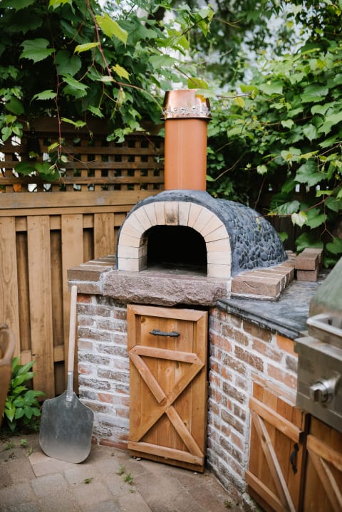 An outdoor pizza oven with a stone and brick structure, copper chimney, and surrounded by plants.