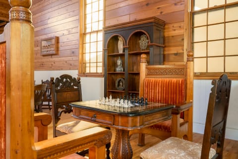 A chess table with clear and black pieces surrounded by ornate wooden chairs in a warmly lit room.