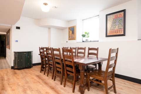 A wooden dining table with chairs in a bright room, featuring artwork on the walls and a plant by the window.