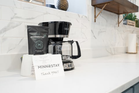 A black coffee maker on a white countertop next to a bag of MINNESTAY coffee and a thank-you card.