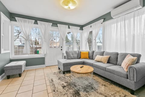 A sunroom featuring a grey sectional couch, wooden coffee table, and large windows with white curtains.
