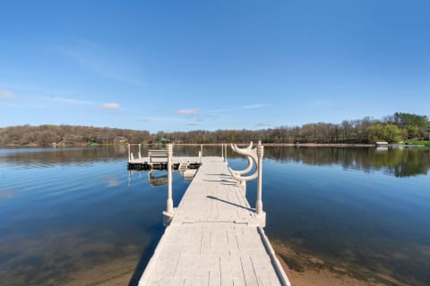 A long dock extending over calm waters, with trees and houses in the background under a bright blue sky.