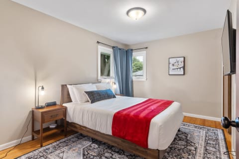 A well-decorated bedroom featuring a bed with white linens and a red throw, a nightstand with a lamp, and artwork on the wall.