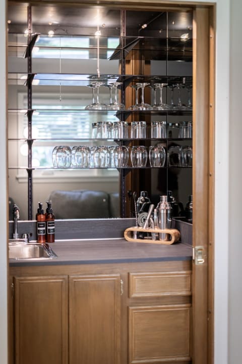 An elegant home bar featuring glass shelves filled with drinkware, a polished sink, and bar tools.