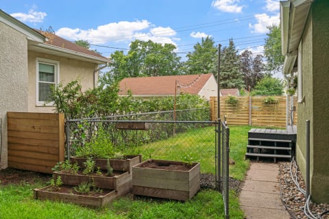 A backyard featuring raised garden beds, a gravel path, and a wooden deck, with trees and a clear blue sky in the background.