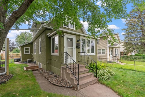 Front view of a green house with stairs, surrounded by a lawn and trees.