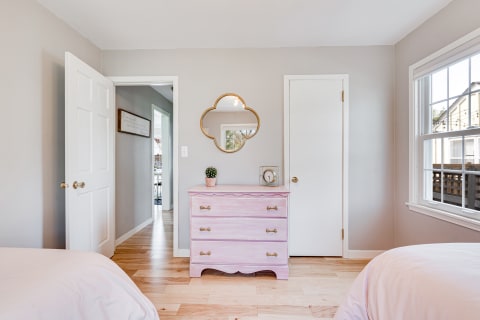 A cozy bedroom featuring a pastel pink dresser, a decorative mirror, and natural light coming through a window.