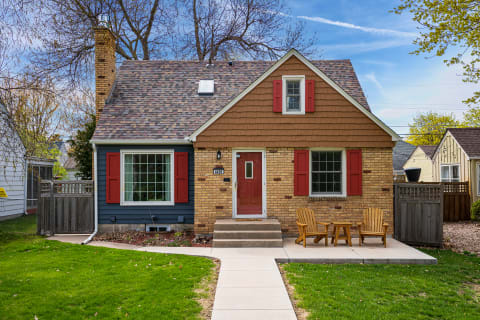 A cozy house with a steep roof, red shutters, and wooden chairs in the yard.
