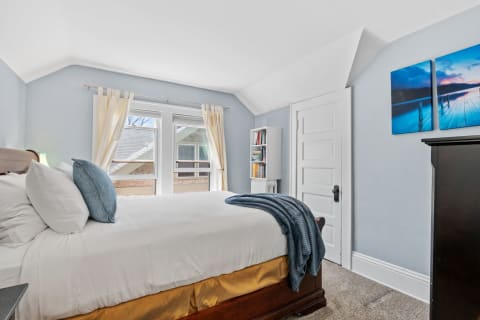 A peaceful bedroom featuring a bed with white linens and a blue throw blanket, light blue walls, natural light from windows, and a bookshelf.