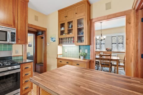 A warm kitchen with wooden cabinets, green tiled backsplash, and a dining area visible through an open door.