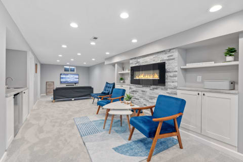 Cozy modern basement living area with blue chairs, a fireplace, and a kitchenette.